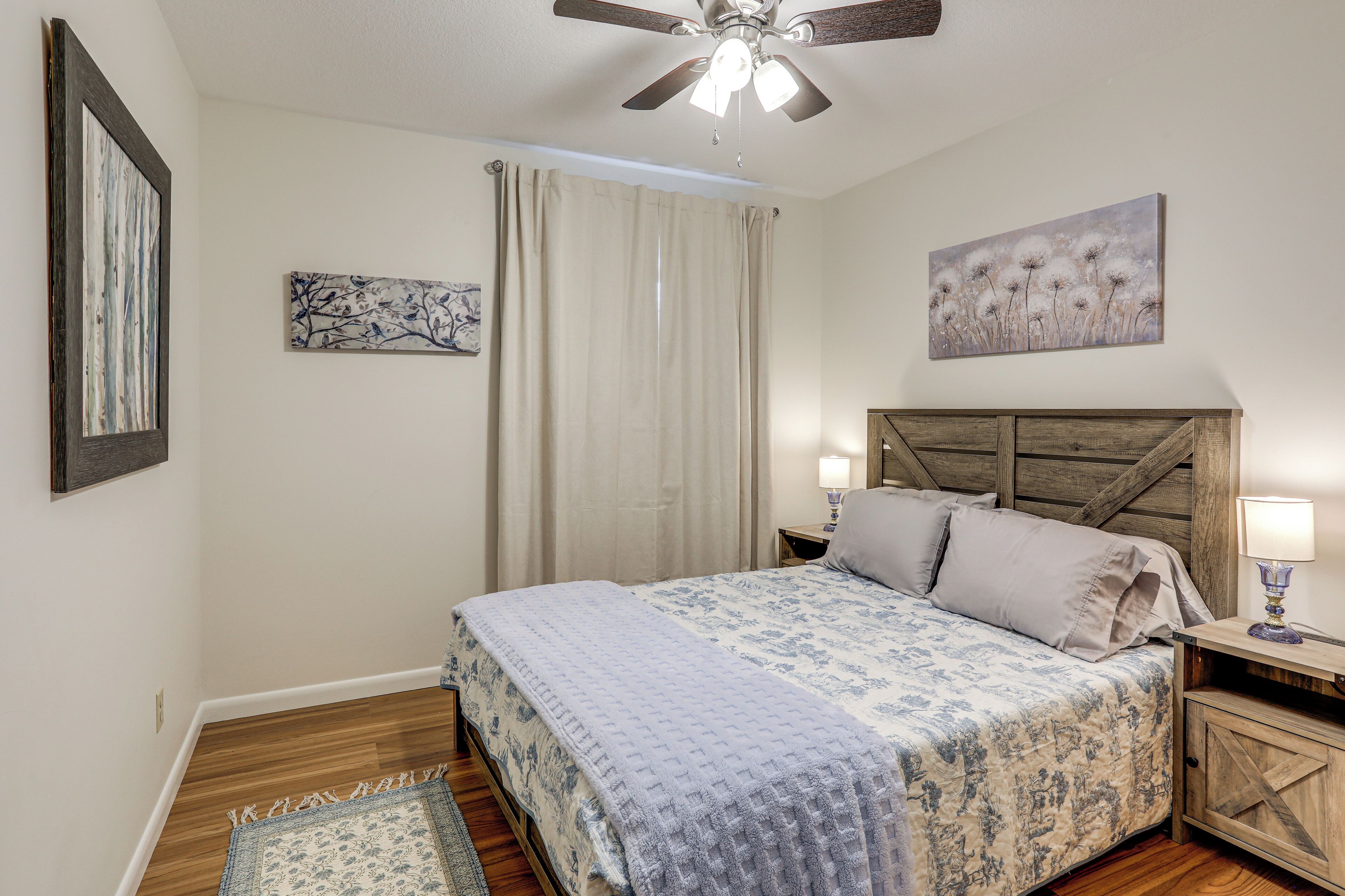 A bedroom with a bed, a ceiling fan, a nightstand, a lamp, and artwork on the walls.