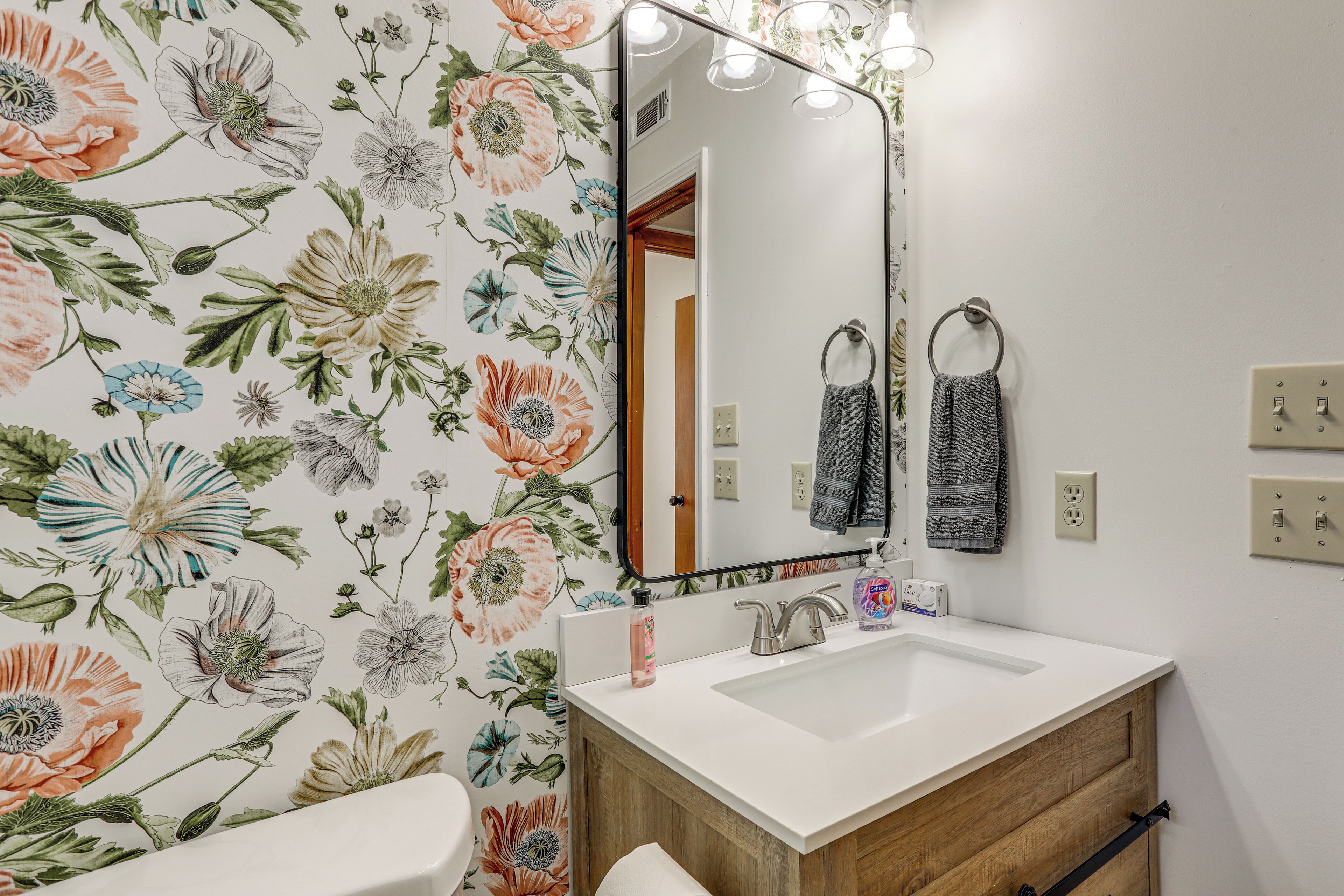 A bathroom with a floral wallpaper, a white sink, a mirror, and wooden cabinets.