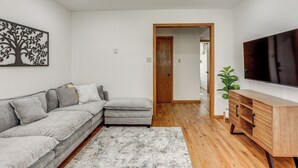 Un salon avec un canapé d’angle gris, un meuble en bois, un téléviseur à écran plat et une porte donnant sur une autre pièce.