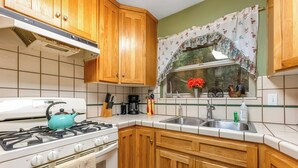 A kitchen with wooden cabinets, a stove, a sink, and a window with a floral curtain. - Rustic Mtn-view Home w/ Wood Stove: Dog Friendly (Felton)