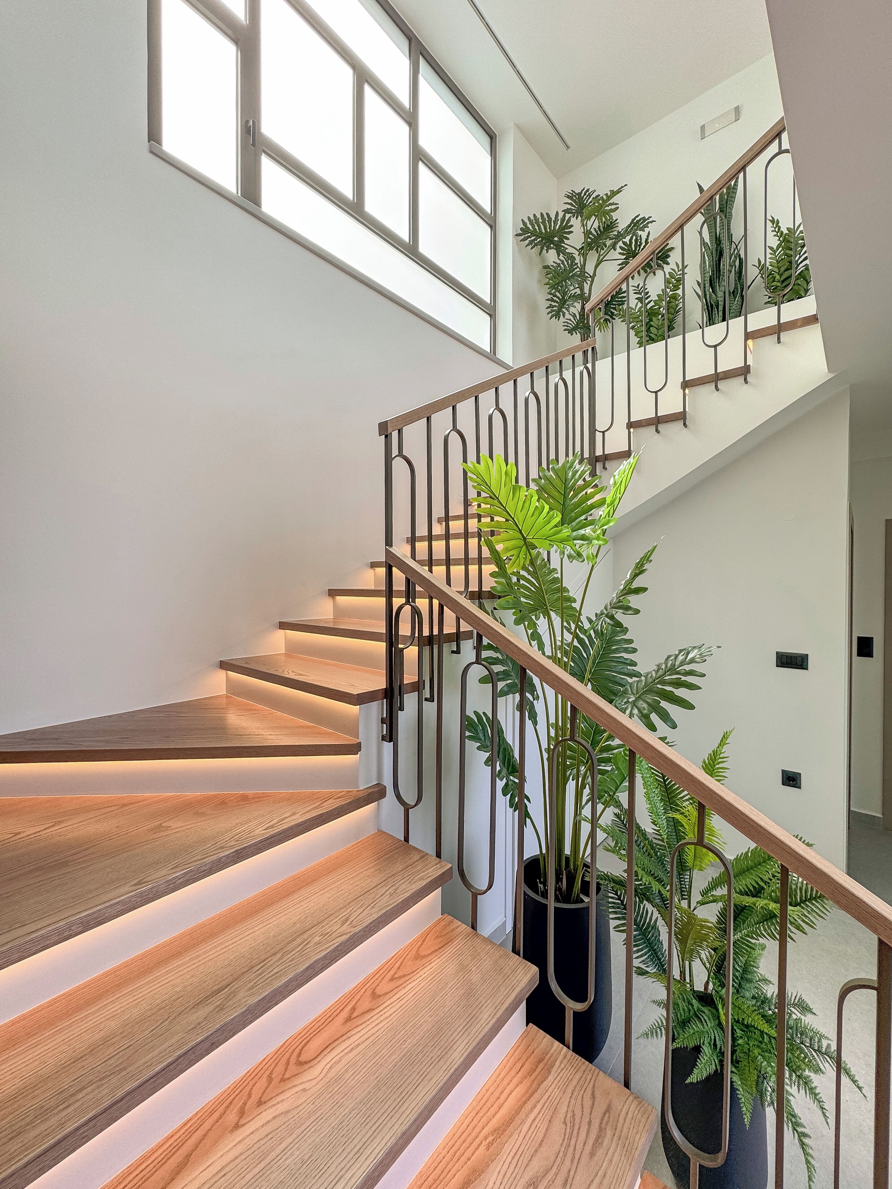 Una escalera moderna con peldaños de madera y barandillas de metal, rodeada de plantas en macetas.