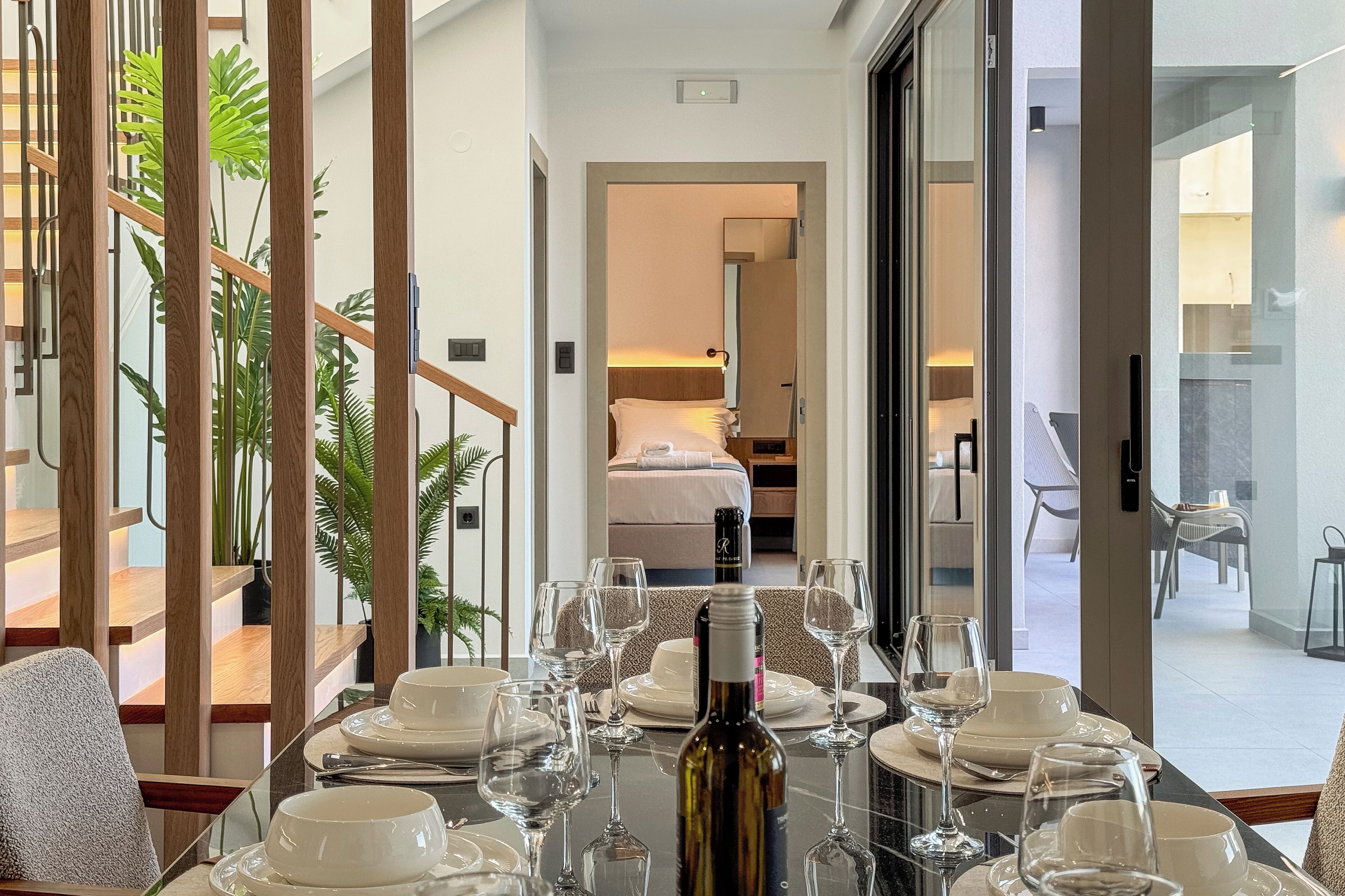Un comedor con una mesa dispuesta para una comida, una botella de vino y vistas a un dormitorio con cama y mesita de noche.