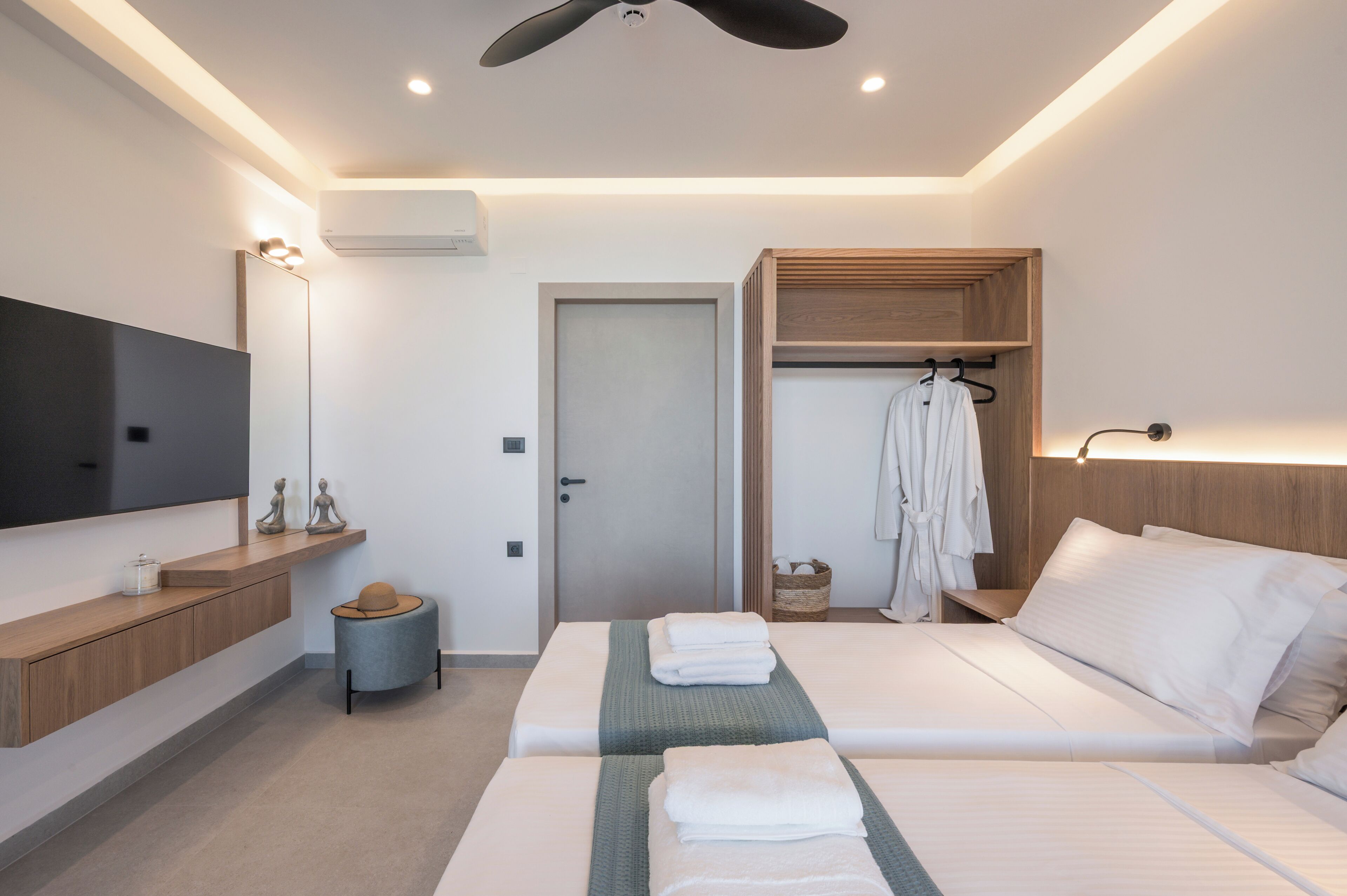 A modern hotel room with a flat-screen TV, a wooden vanity, a ceiling fan, a bed with white linens, and a robe hanging on a wooden rack.