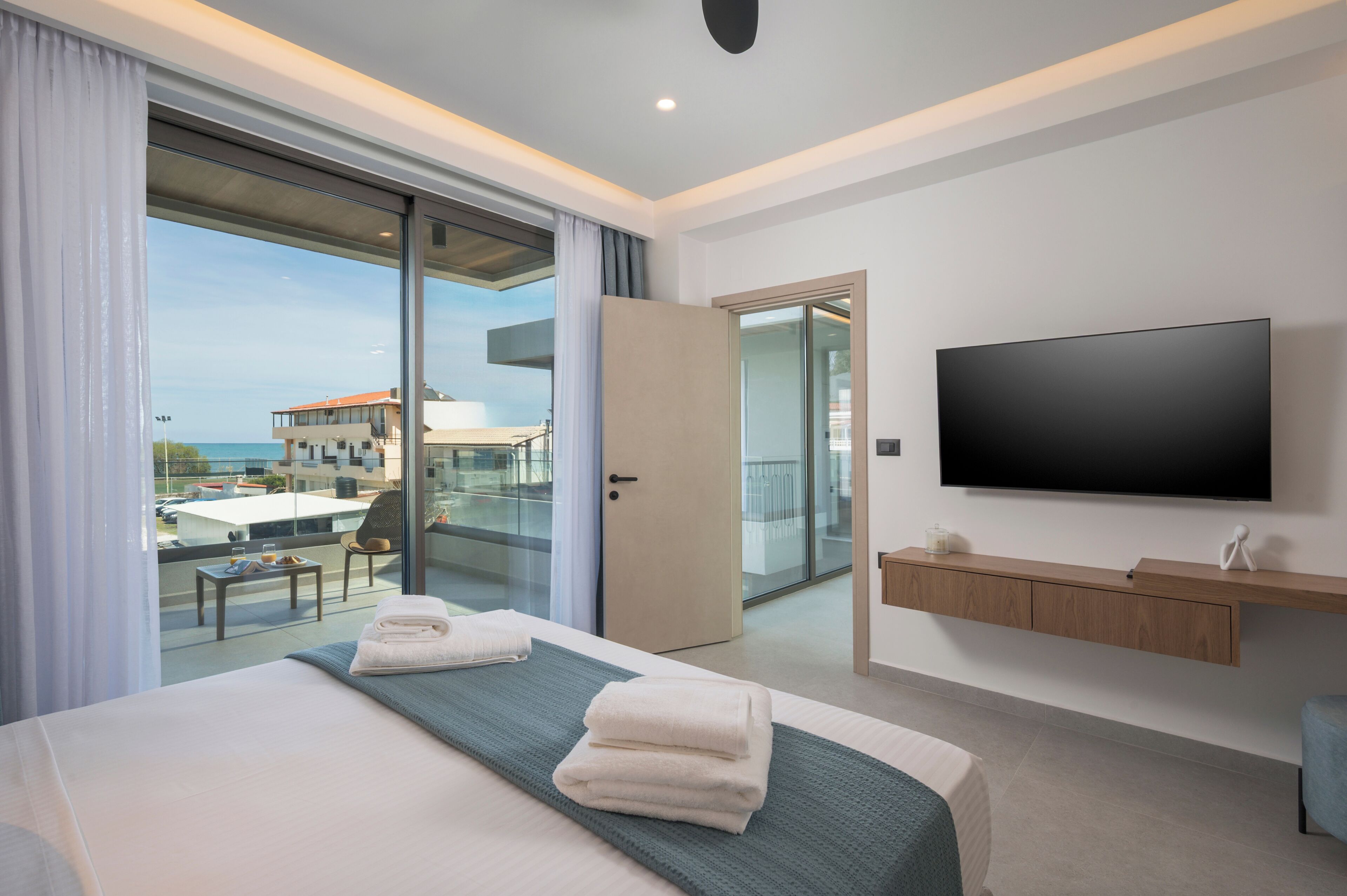 A modern hotel room with a large bed, a TV, a balcony with a view, and a sliding glass door.