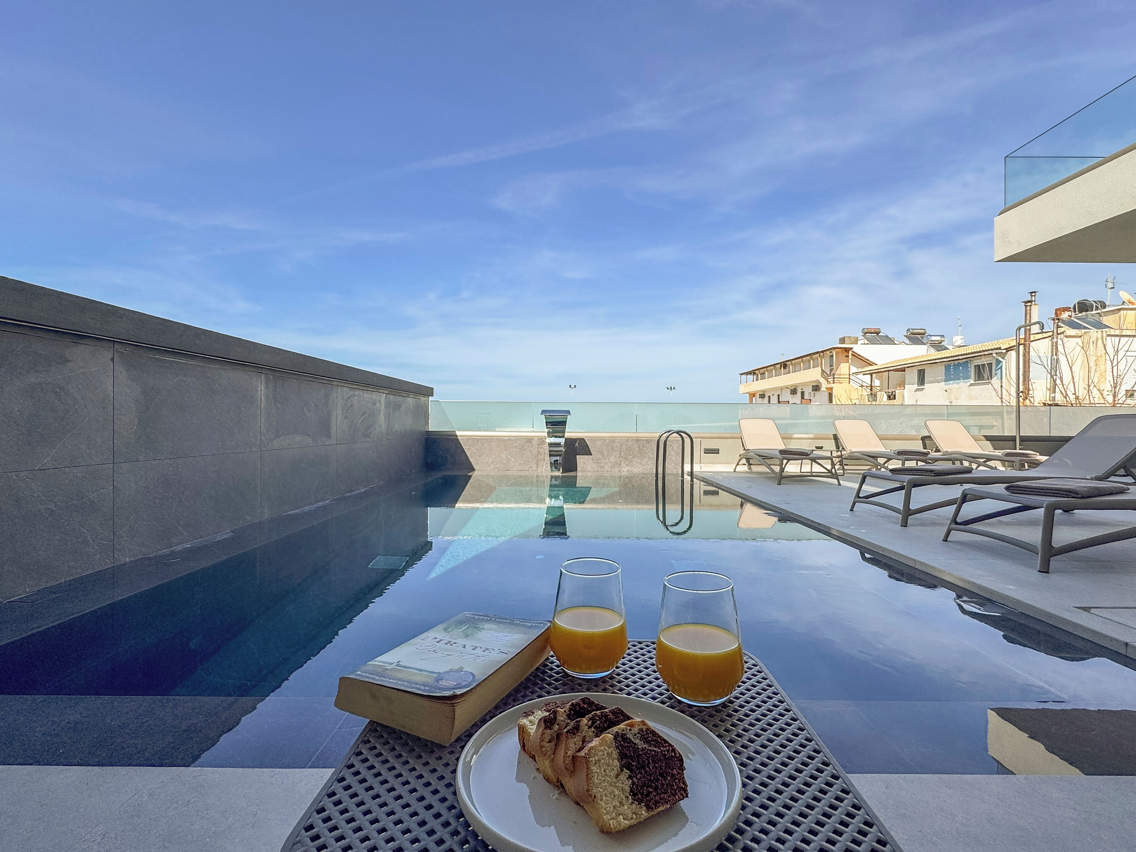 A poolside table with a book, two glasses of orange juice, and a plate with a slice of cake.
