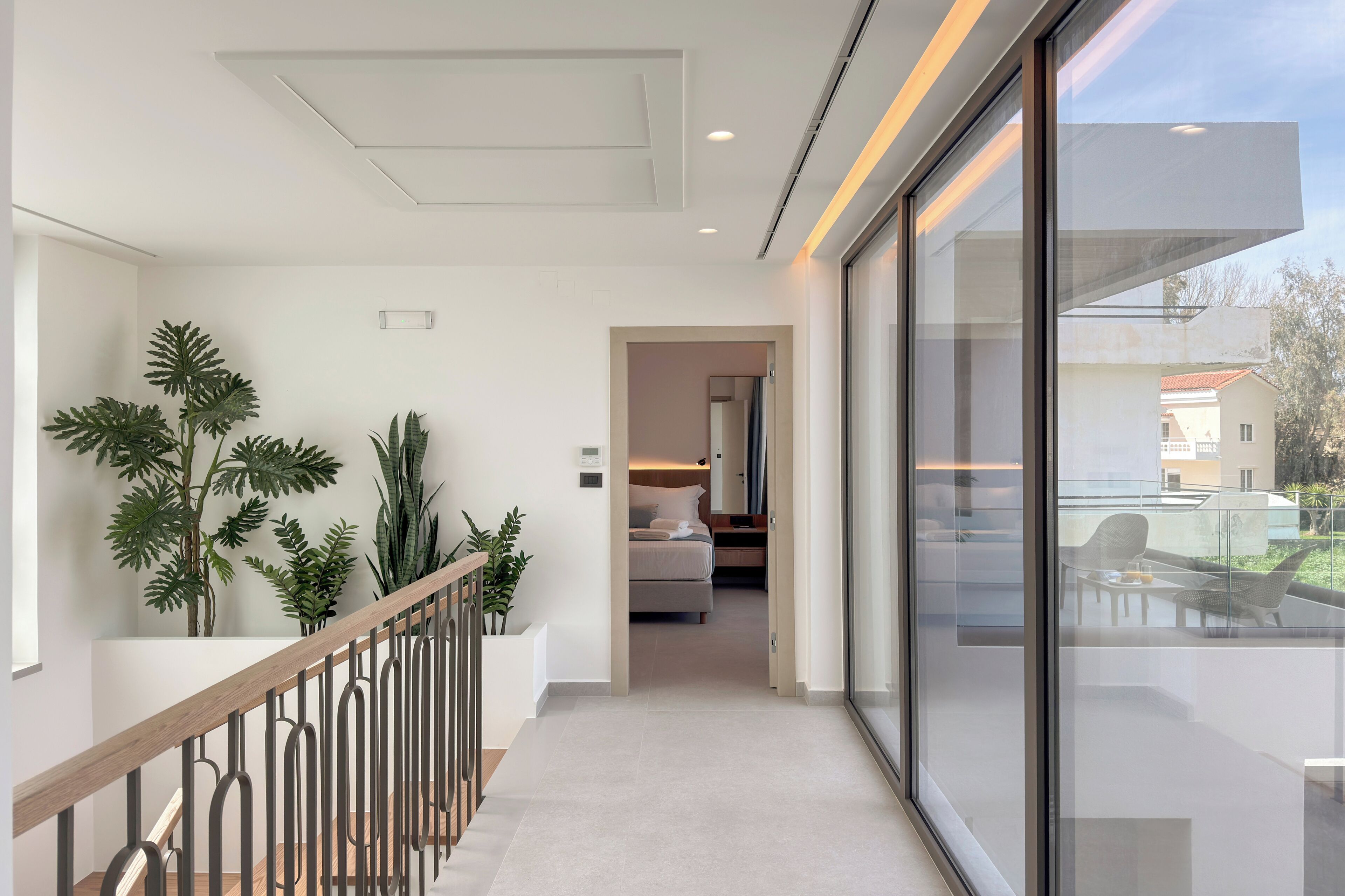 A modern hotel interior with a staircase, potted plants, a bedroom with a bed and nightstand, and a balcony with outdoor furniture.