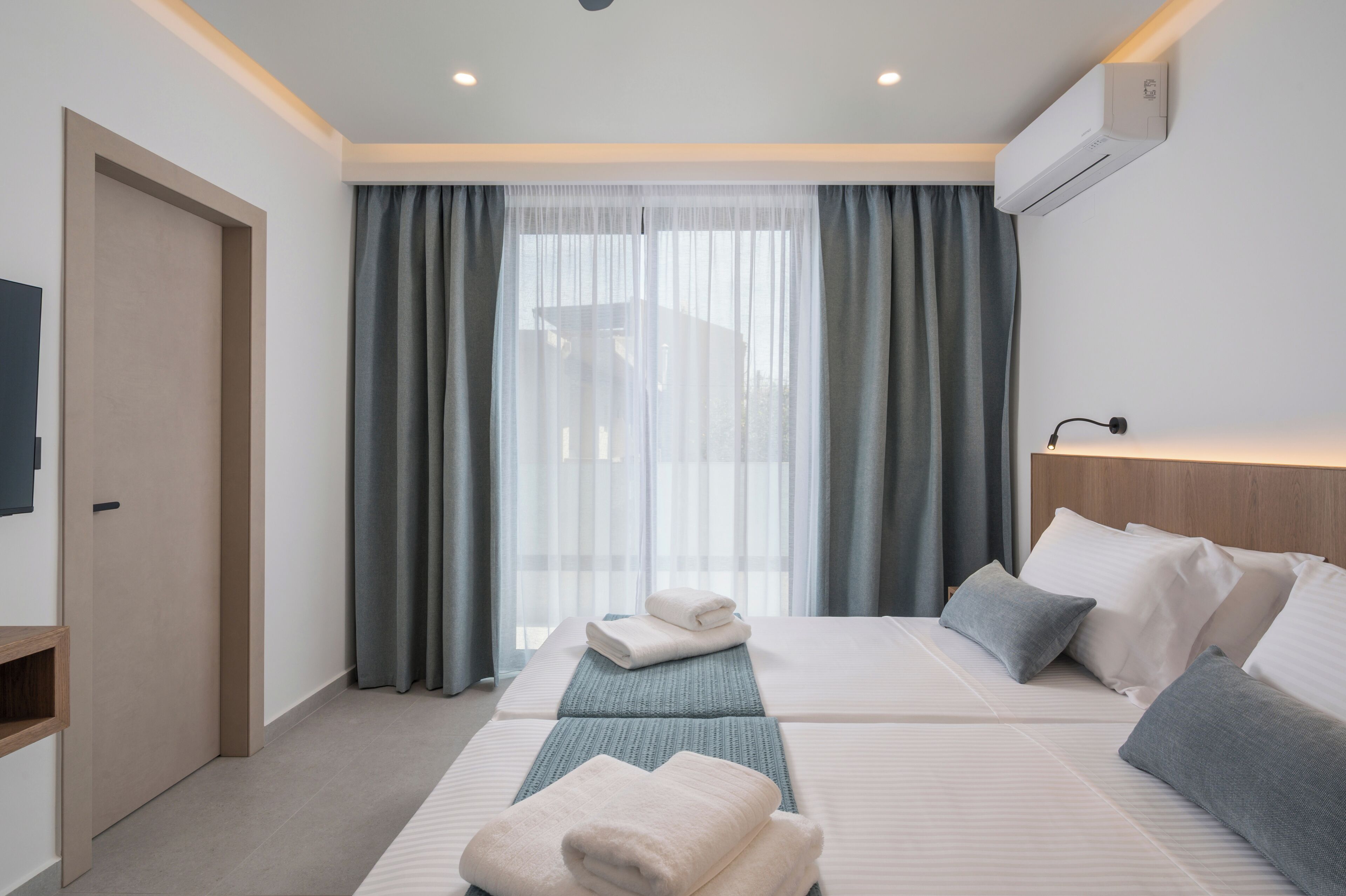 A hotel room with two beds, a TV, an air conditioner, and a view of the outside.
