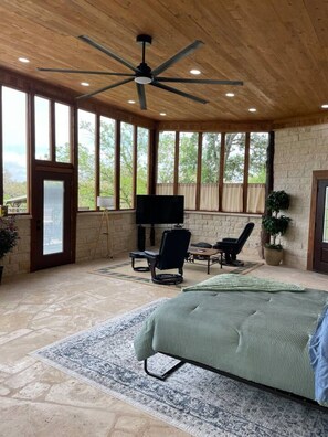 A spacious room with a large bed, a ceiling fan, a TV, and a stone wall. - The Cross Patio Retreat - Designer Hill Country Studio with Scenic Views (Buda)