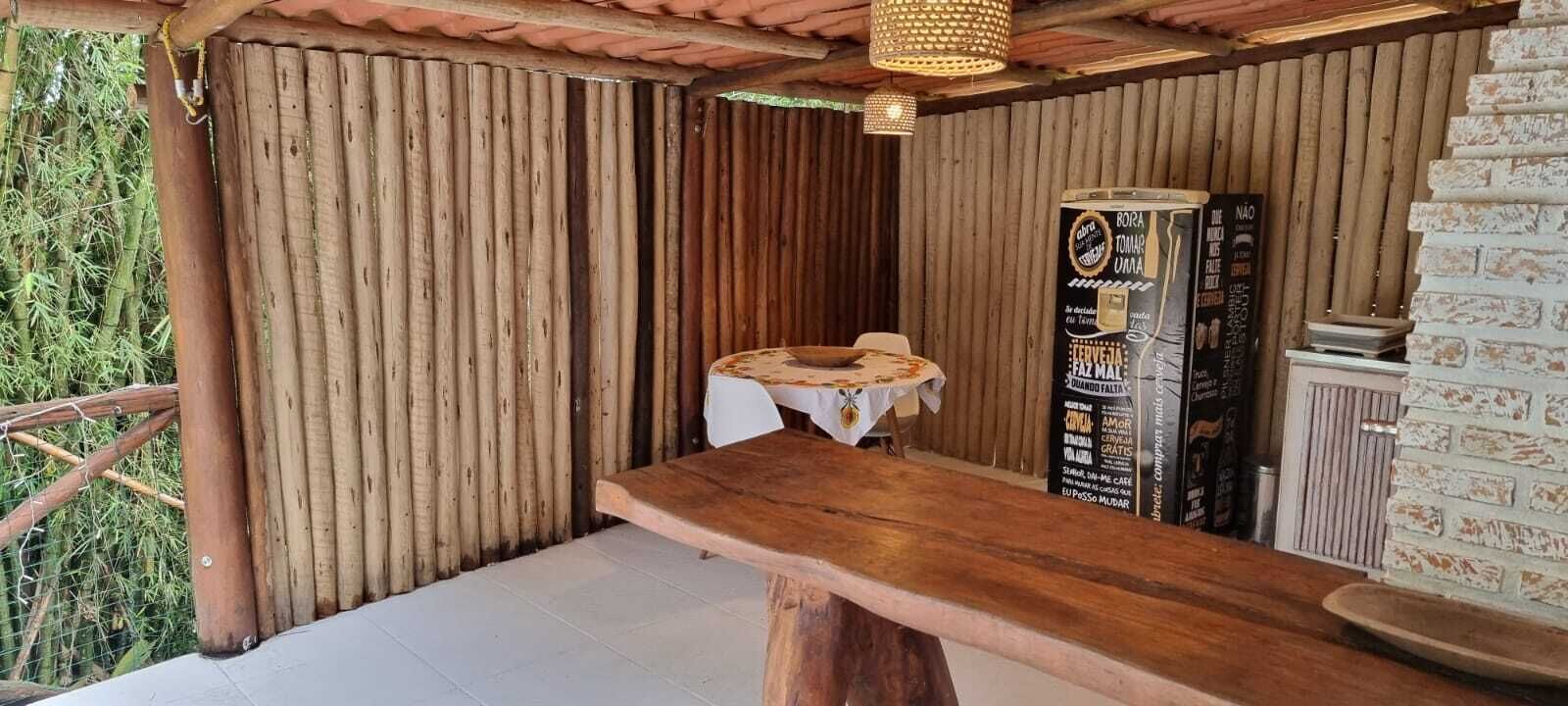 Una mesa de madera rústica con mantel blanco, un taburete de madera y un panel con el menú en un espacio al aire libre cubierto, con paredes de bambú y una pared de ladrillo.