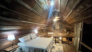 A cozy, rustic bedroom with a bed, bedside lamp, wooden walls, and a hanging light fixture.