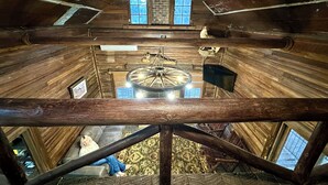 A wooden interior with a central chandelier, a brick fireplace, and a view of a person sitting on a couch.