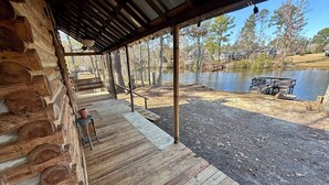 A wooden cabin with a porch overlooking a lake, a wooden deck, and a bench.