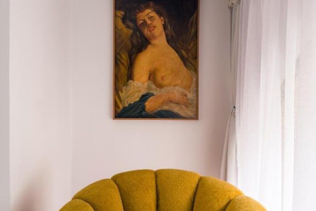 A room with a painting on the wall, a yellow armchair, and a white curtain.