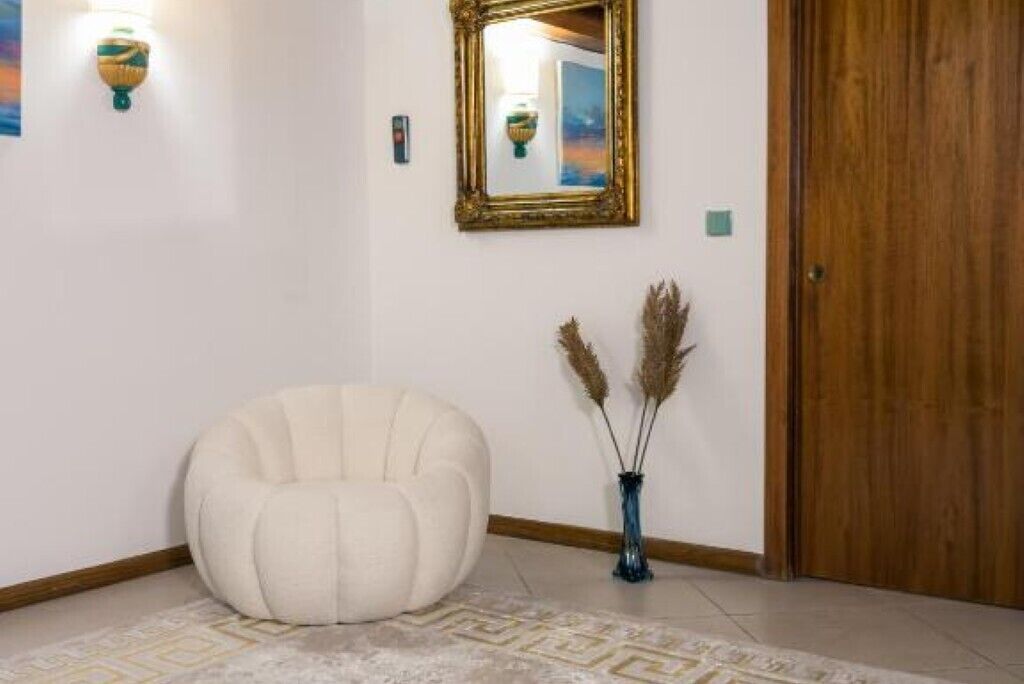 Ein Zimmer mit einem runden, cremefarbenen Sessel, einem Spiegel mit goldener Rahmung, einer Vase mit Trockenpflanzen und einer Holztür.