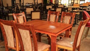 Une table à manger en bois, entourée de six chaises, et disposée dans une pièce où se trouvent d’autres tables et chaises, le sol étant recouvert de carreaux.