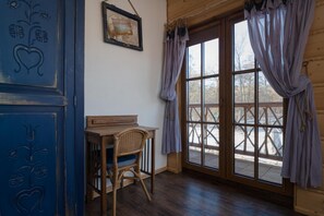 Ein Zimmer mit einem Holztisch, einem Korbstuhl, einem großen Fenster mit Vorhängen und einer Wand mit dekorativen Schnitzereien.