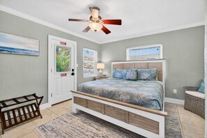 A bedroom with a bed, a ceiling fan, a door, a window, a nightstand, a crib, and a wicker chair. - Coconut Villa`s - Suite 10 (Redington Shores)