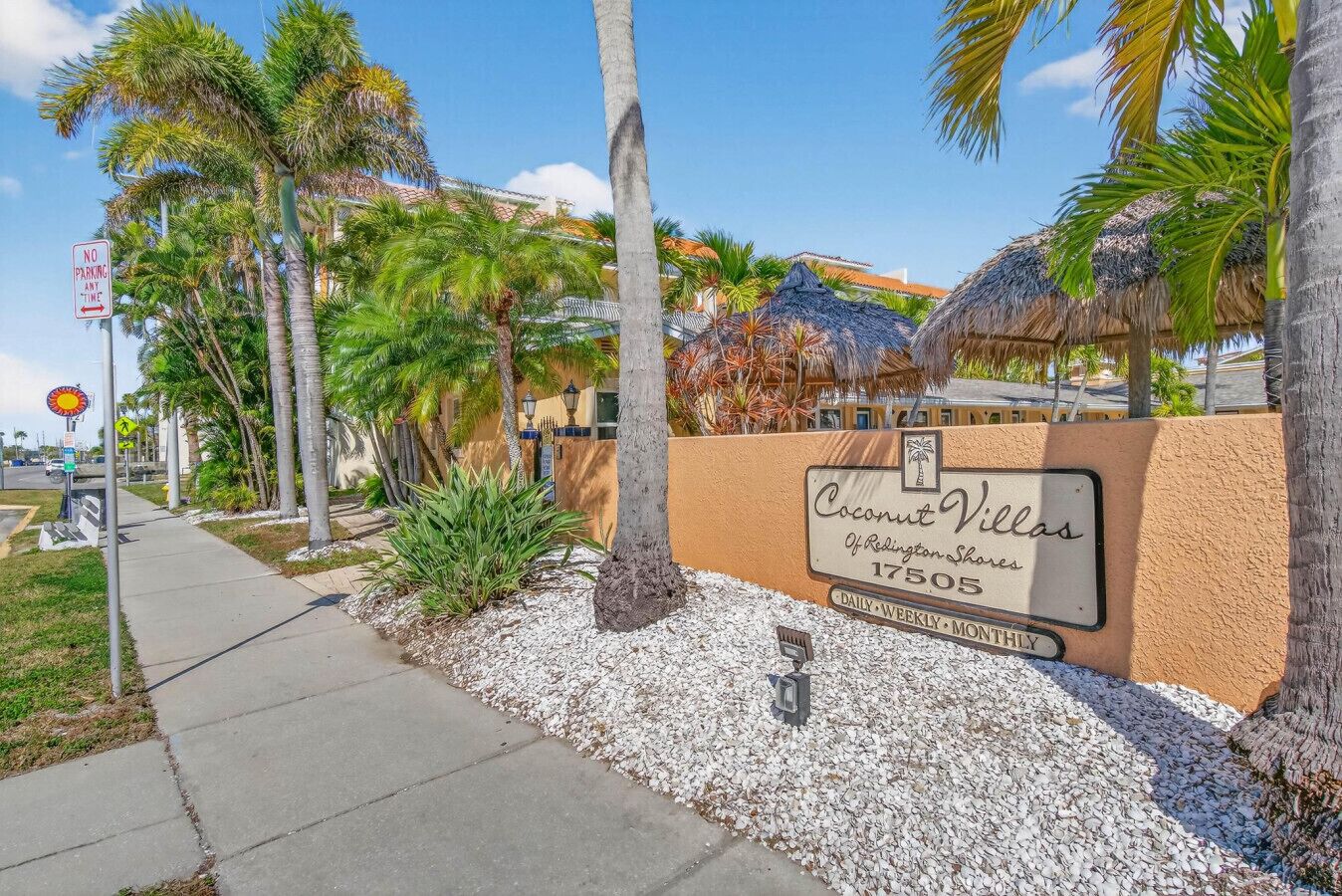 Una señal de calle indica Coconut Villas, un edificio con entrada bordeada de palmeras y cielo despejado.