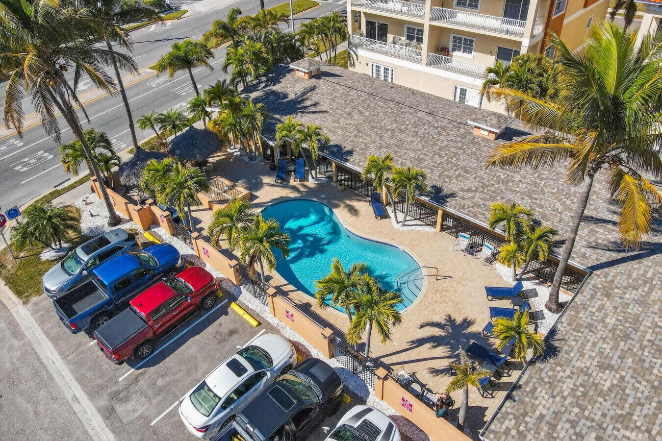 Vista aérea de un hotel con piscina, coches estacionados y palmeras.