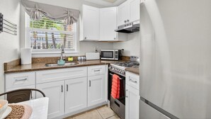A kitchen with white cabinets, a microwave, a stove, a refrigerator, a window with curtains, and a sink. - Coconut Villas Suite 5 (Redington Shores)