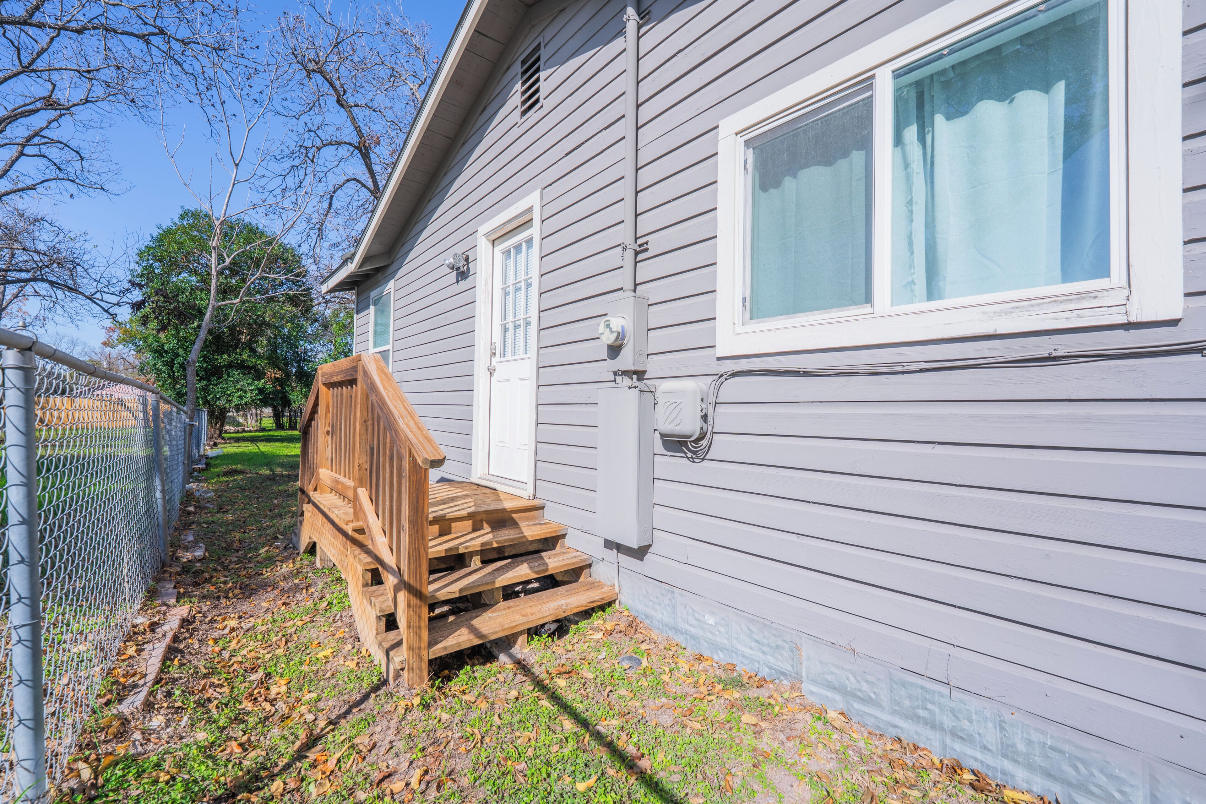 A house with a wooden staircase leading to a white door, a window with a curtain, and a chain-link fence.