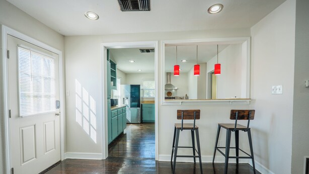 Un pasillo con una puerta, una zona de cocina con armarios azul verdosos y dos taburetes.