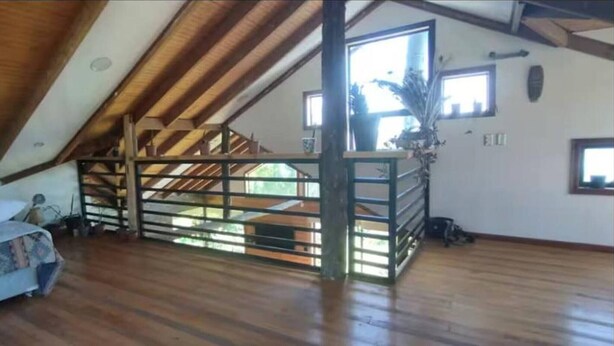 A loft-style room with a wooden ceiling, a bed with patterned bedding, a staircase, and a large window.