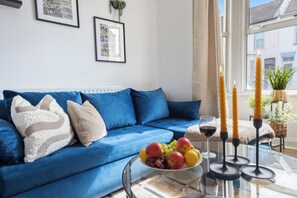 A blue sofa with decorative pillows, a glass table with a fruit bowl, candles, and a wine glass. - Stylish and Cosy 3Bd- WiFi- Long Stay Ready (Gillingham)