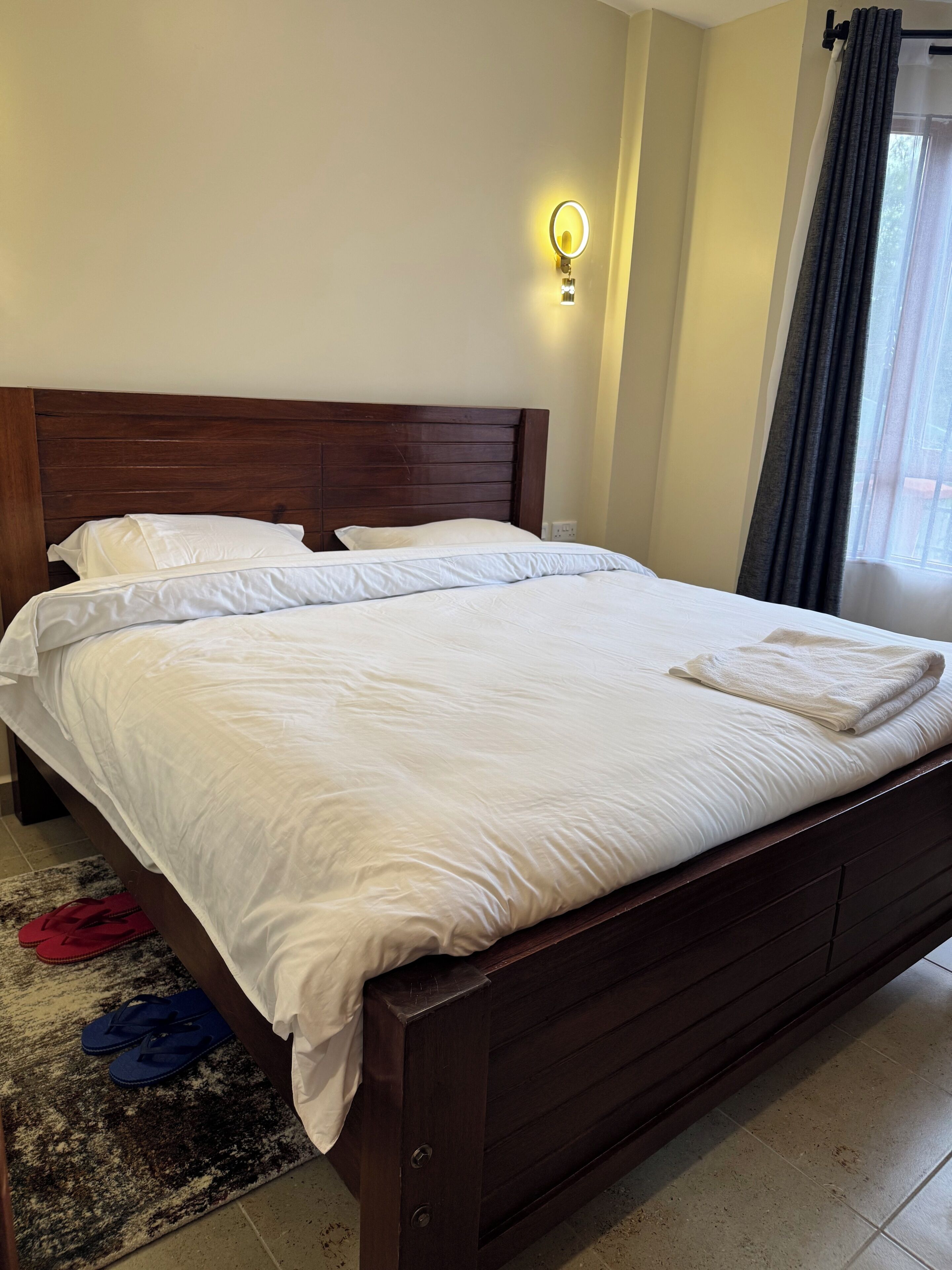 Una cama bien hecha con sábanas blancas, cabecera de madera y una lámpara de pared pequeña.