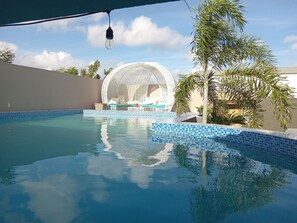 A swimming pool with a clear blue water surface, a white gazebo-like structure, and a palm tree.