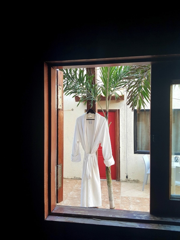 A window view of a room with a white robe hanging on a rack, a red door, and a small tree.