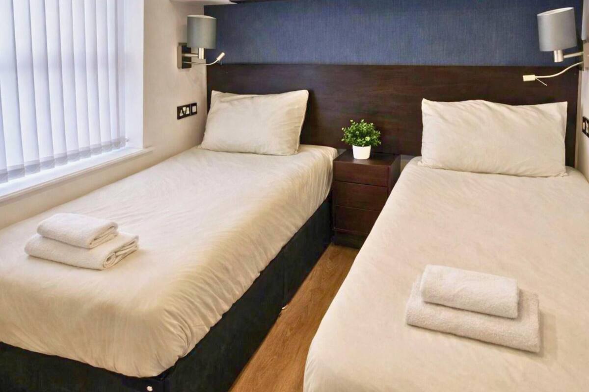 A hotel room with two beds, each with a pillow and a folded towel. There is a nightstand with a lamp and a small plant.