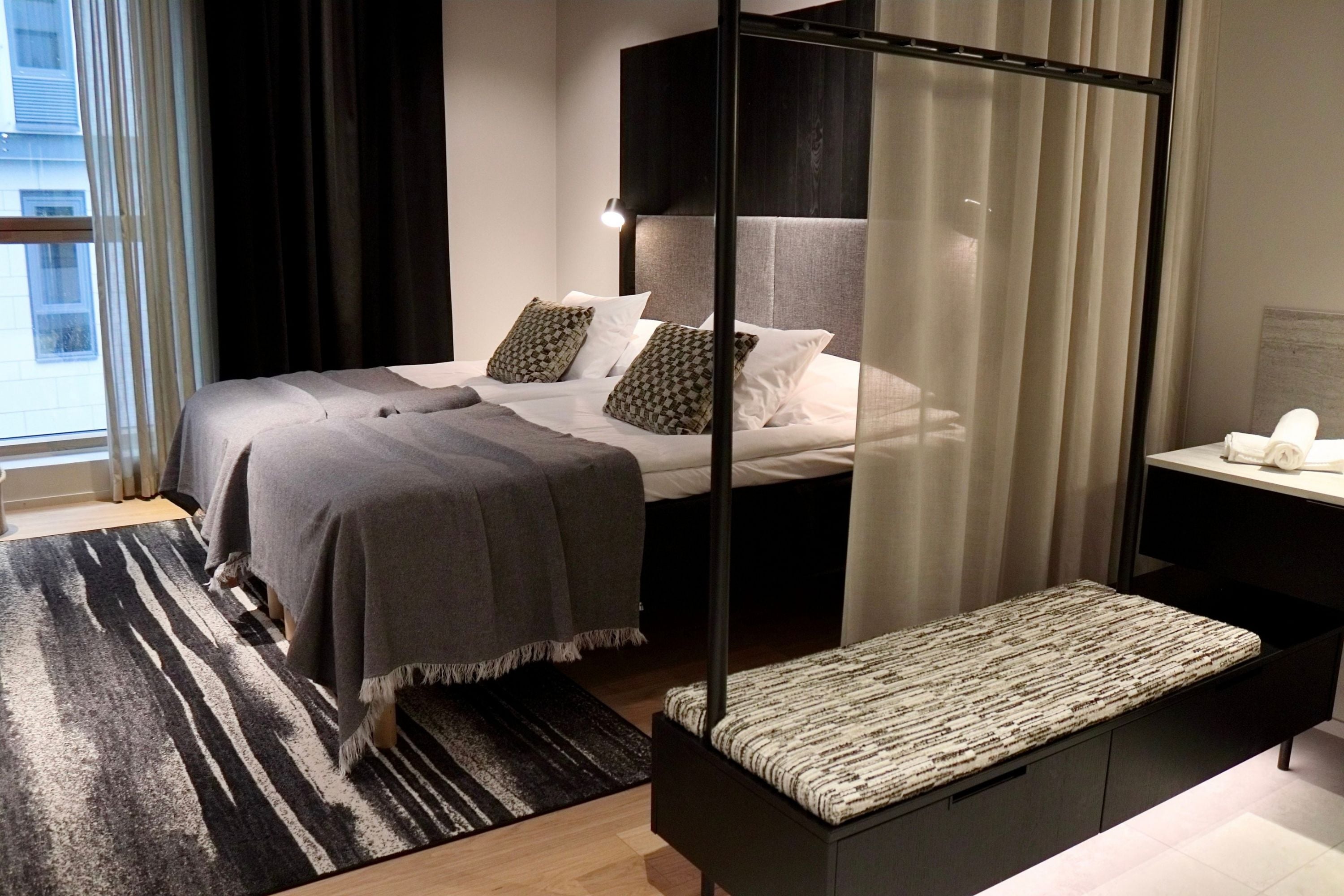 Un dormitorio moderno con cama, mesitas de noche, un banco y vistas al exterior.