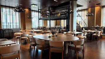 A spacious restaurant with high ceilings, wooden beams, and large windows. Tables are arranged in a casual layout, and there are multiple seating options including booths and chairs.