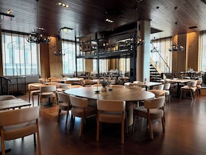 Un restaurant spacieux avec de hauts plafonds, des poutres en bois et de grandes fenêtres. Les tables sont disposées de manière informelle, et différents types de sièges sont proposés, notamment des banquettes et des chaises.