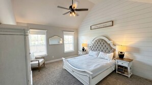 Une chambre avec un grand lit, un ventilateur de plafond, deux lampes de chevet, une table de chevet, une chaise et une fenêtre avec des stores.