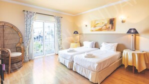 A hotel room with two beds, a wicker chair, a large window with curtains, and a painting on the wall. - VILLA IRLANDAIS (Alvor)
