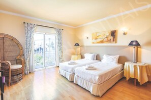 Ein Hotelzimmer mit zwei Betten, einem Korbstuhl, einem großen Fenster mit Vorhängen und einem Gemälde an der Wand.