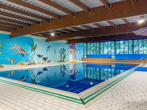 Une piscine intérieure dont les murs sont ornés d’une fresque représentant la vie marine, avec un plongeoir et de grandes fenêtres offrant une vue dégagée sur l’extérieur.