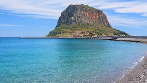 A coastal landscape with a prominent rocky hill, a pier extending into the sea, and a small settlement with buildings. - Matroni s House - Seafront Getaway in Monemvasia (Monemvasia)