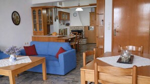 A living room with a blue sofa, wooden dining table, and chairs, a kitchen area with cabinets and a fireplace, and a clock on the wall. - Matroni s House - Seafront Getaway in Monemvasia (Monemvasia)
