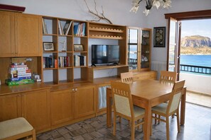 A room with a wooden TV stand, bookshelves, a dining table with chairs, and a view of the sea. - Matroni s House - Seafront Getaway in Monemvasia (Monemvasia)