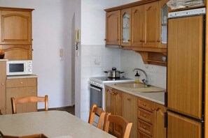 A kitchen with wooden cabinets, a microwave, stove, sink, and chairs around a table. - Matroni s House - Seafront Getaway in Monemvasia (Monemvasia)