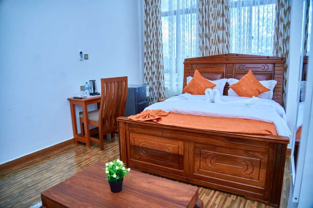 A wooden bed with orange pillows, a small table with a plant, and a chair in a room with patterned curtains and a window.