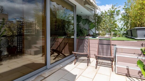 Un patio con dos sillones de mimbre, una puerta de cristal y vistas a un jardín con diversas plantas.