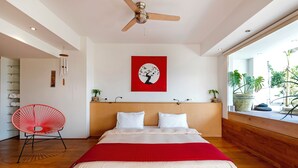 Un dormitorio moderno con una cama, una silla roja, un ventilador de techo y una ventana con plantas.