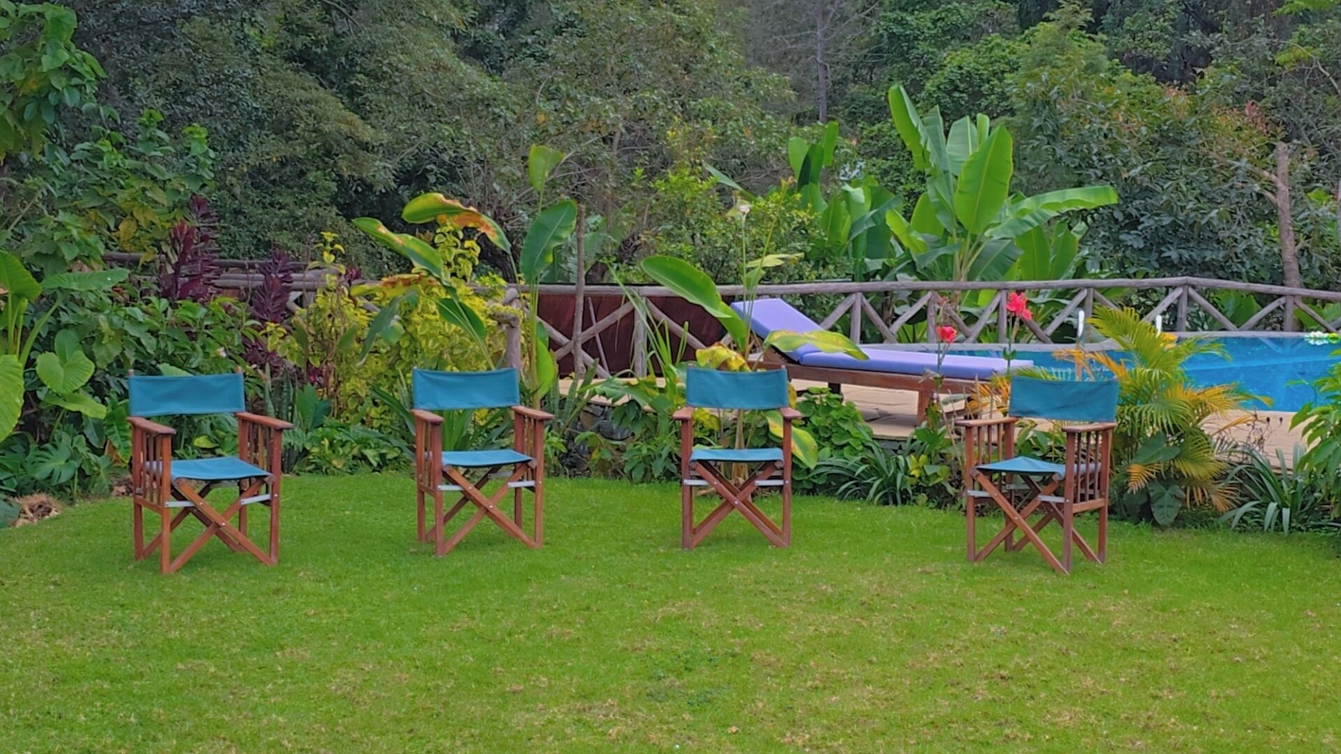 Un espace extérieur aménagé avec quatre chaises en bois et une table bleue, entouré d’une végétation luxuriante et d’une clôture en bois.