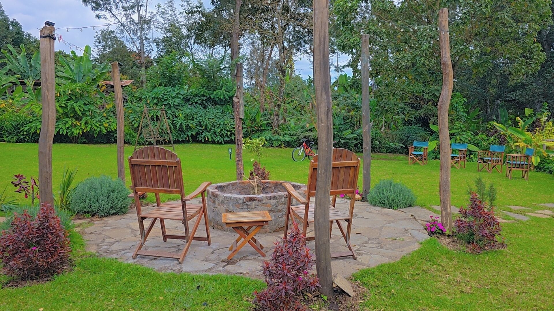 An outdoor seating area with wooden chairs and a stone table, surrounded by a lush garden and trees.