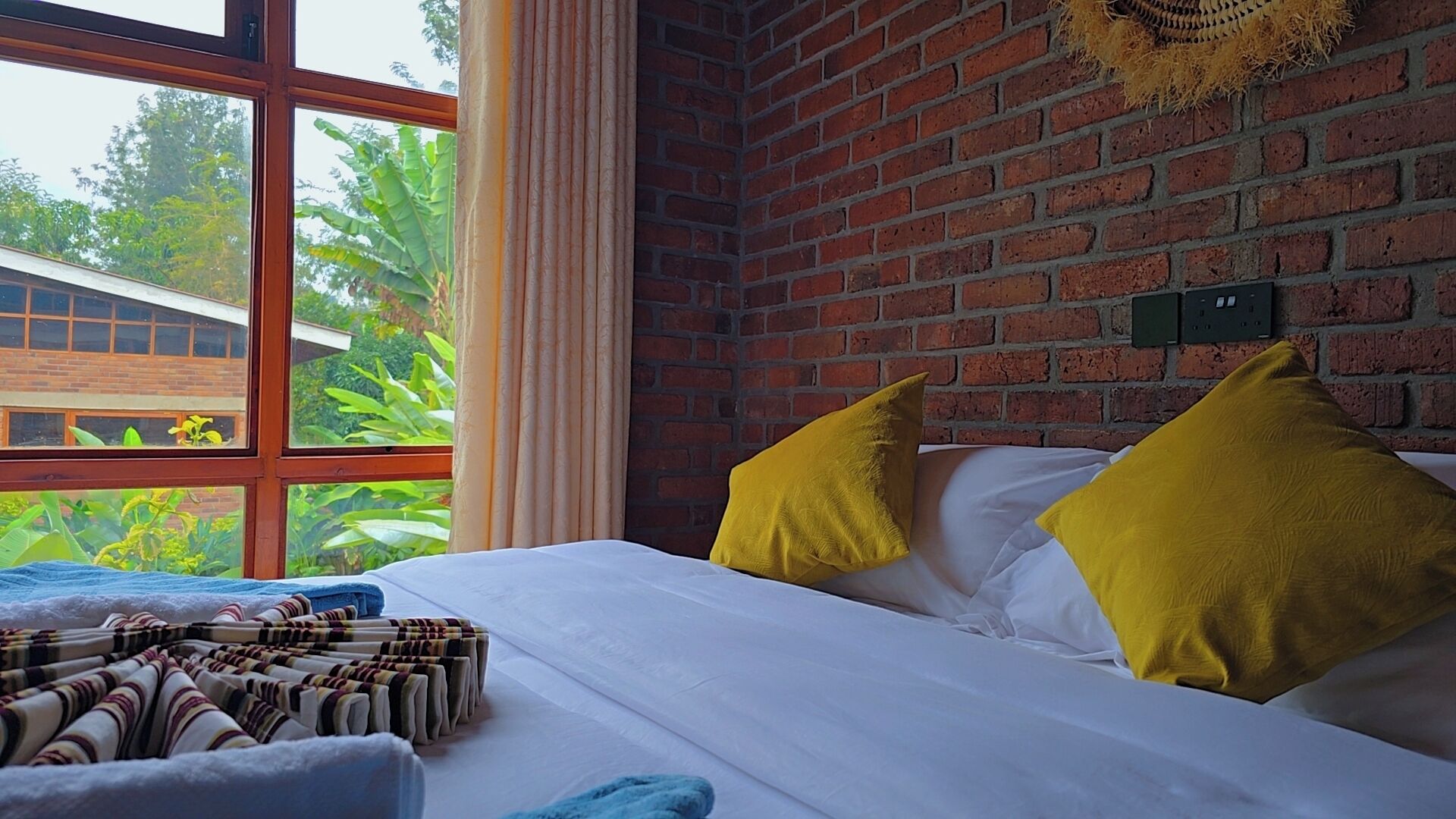 A room with a brick wall, a bed with white bedding and two yellow pillows, a window with curtains, and a view of greenery outside.