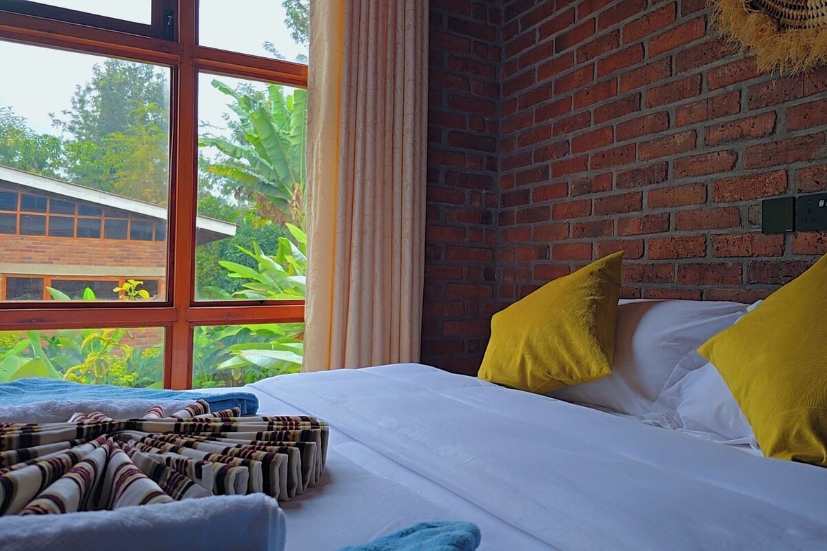 A room with a brick wall, a bed with white bedding and two yellow pillows, a window with curtains, and a view of greenery outside.